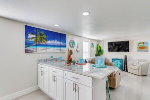 A modern kitchen with white cabinets, a marble countertop, and a large island. There is a painting of a beach scene on the wall, a clock, and a guitar leaning against the wall. - Gulf Breeze Home w/ Deck: Walk to Public Boat Ramp (Gulf Breeze)