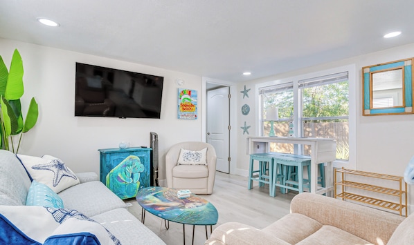 A living room with a sofa, a coffee table, a TV, a chair, and a dining area with a table and chairs. - Gulf Breeze Home w/ Deck: Walk to Public Boat Ramp (Gulf Breeze)