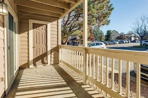 A covered porch with a wooden railing, a door, and a view of parked cars and trees in the background.
