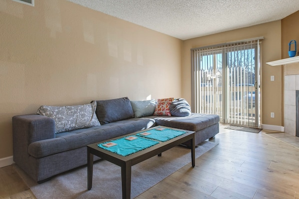 A living room with a grey sofa, a coffee table with placemats, and a sliding glass door leading to a balcony.