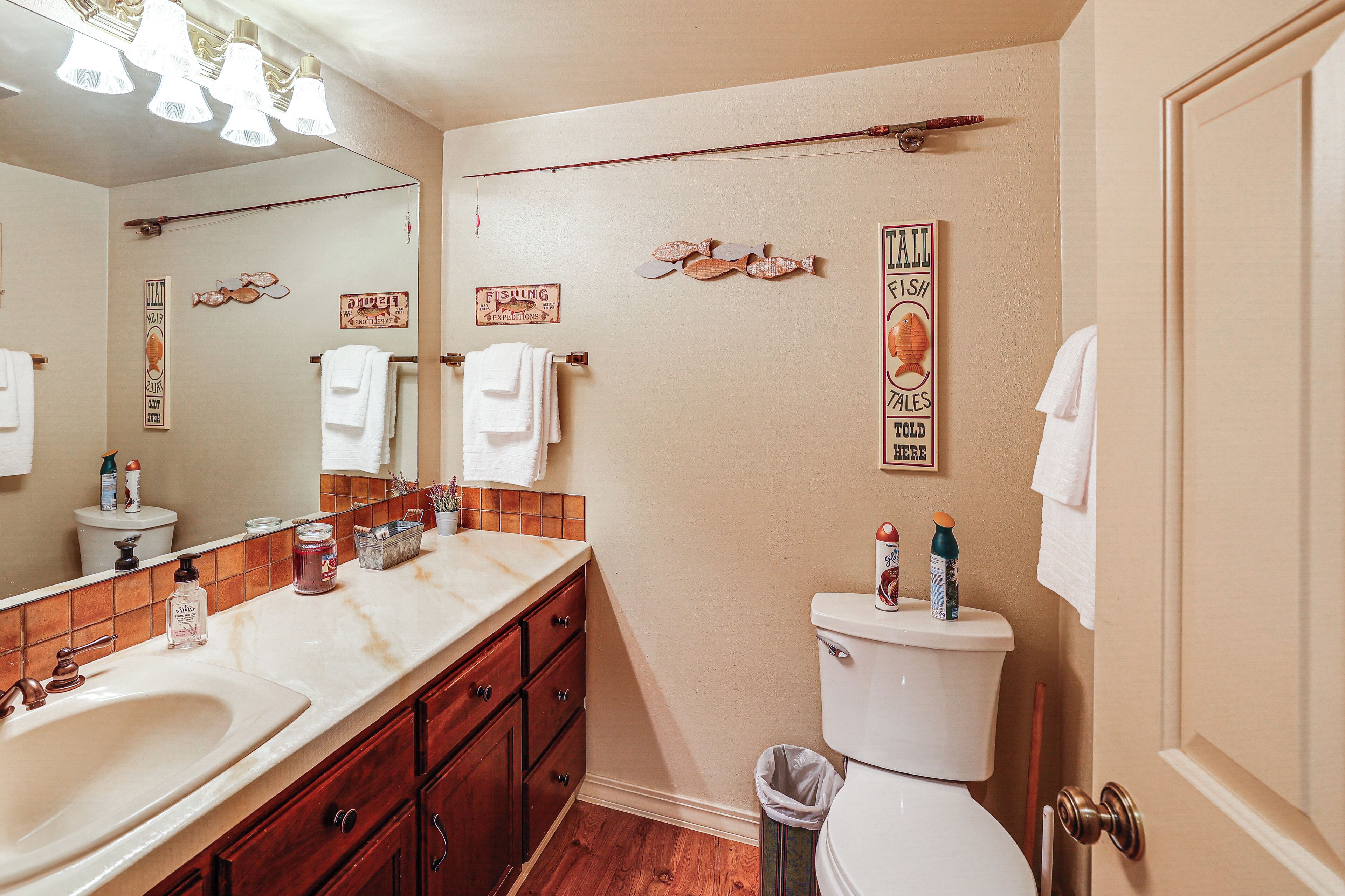 Une salle de bain avec un grand miroir, un lavabo, des toilettes et une poubelle. Des serviettes sont accrochées au mur, et il y a un panneau indiquant un lieu de pêche.