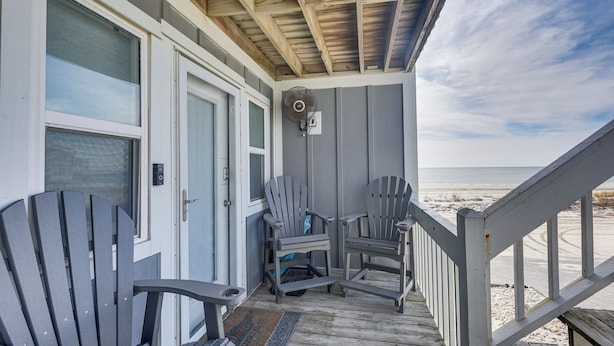 Un porche cubierto con dos sillas Adirondack, una terraza de madera y vista a la playa.