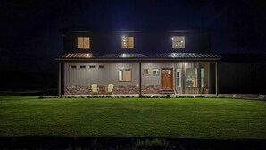 Une maison à deux étages avec une façade en pierre, un porche à l’avant avec des chaises et un éclairage extérieur soigné la nuit.