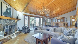 A spacious living room with a stone fireplace, wooden ceiling, and large windows. - Big Bear Lake Cabin w/ Hot Tub & Gas Fire Pit! (Big Bear Lake)