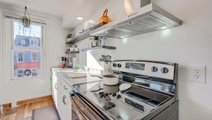 Eine moderne Küche mit einem Edelstahlherd, einem eingebauten Backofen und einer Dunstabzugshaube. Darüber befinden sich Regale mit Küchenutensilien und ein Fenster mit Blick auf umliegende Gebäude.