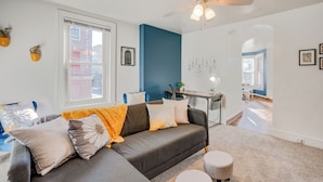 Ein modernes Wohnzimmer mit grauem Ecksofa, Deckenventilator und Blick auf die Stadt durch große Fenster.
