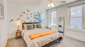 Ein Schlafzimmer mit Bett, Deckenventilator, Nachttisch, Lampe, Fenster und Wandbild.