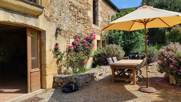 A stone building with a wooden table and chairs under a yellow umbrella, surrounded by gravel and flowering plants. - Agréable Maison en Pierres, à Quelques Minutes des Châteaux (vézac)