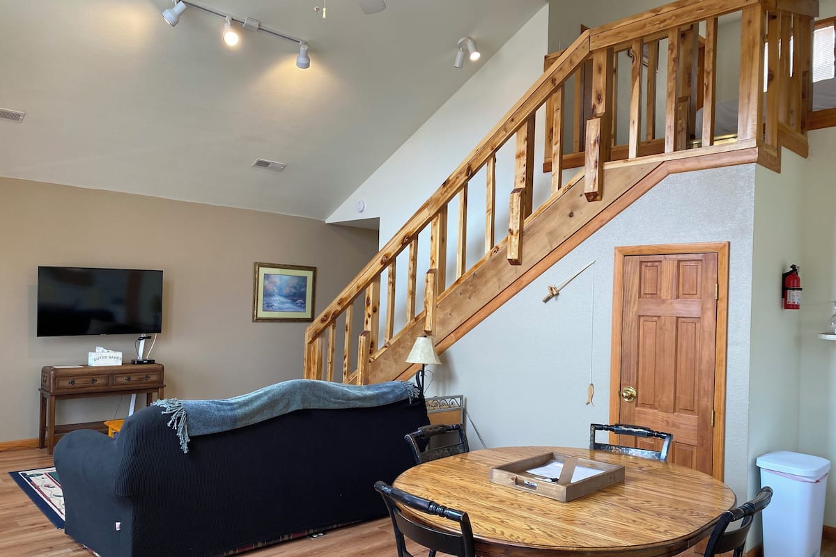 Una sala de estar acogedora con una escalera de madera, un televisor, un cuadro y un comedor con una mesa y sillas.