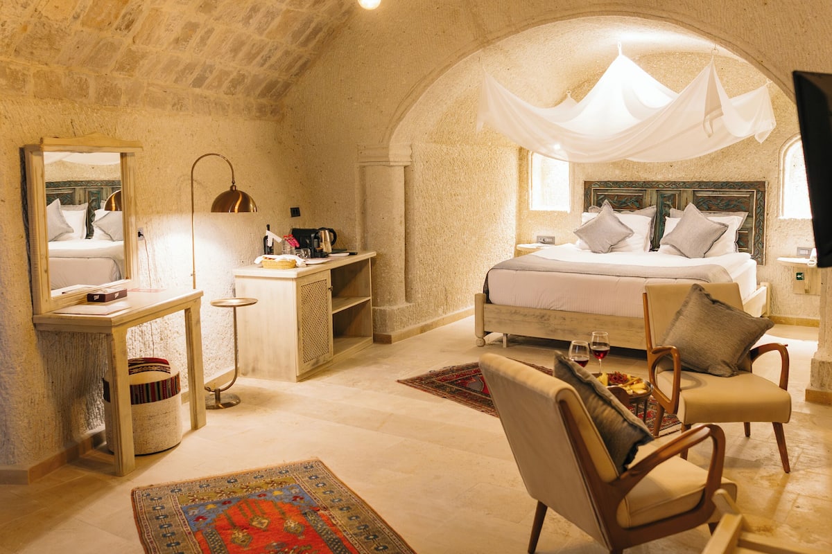 A cozy bedroom with a stone ceiling, a large bed with a canopy, a bedside table, a chair, and a desk with a lamp.