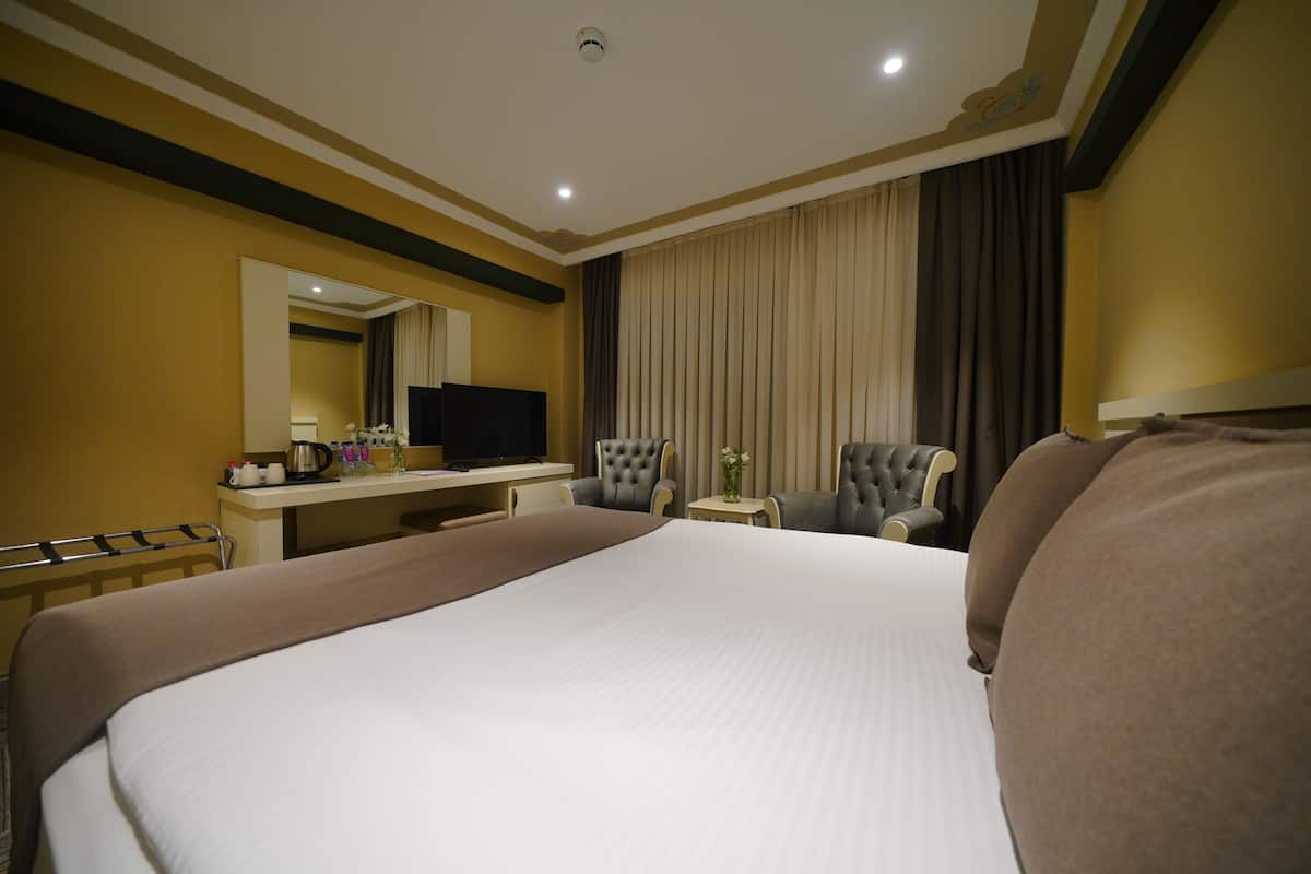 A hotel room with a large bed, a desk with a television, two armchairs, and a small table with a vase.