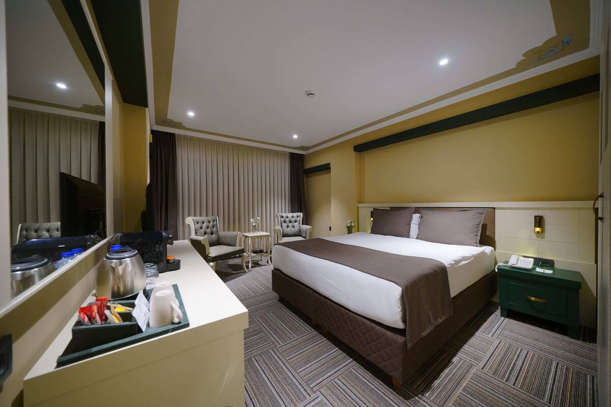 A hotel room with a large bed, a desk with a coffee maker, a chair, a small table, and a nightstand with a lamp.