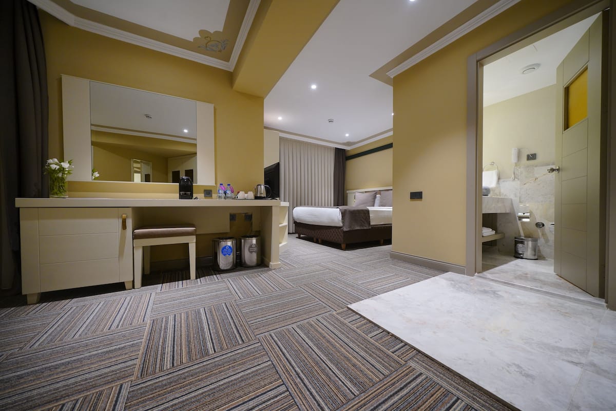 A hotel room with a large bed, a bedside table, a mirror, a bathroom with a toilet and sink, and a carpeted floor.