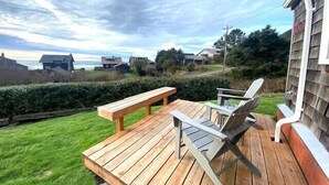 Eine Holzterrasse mit einer Bank und zwei Stühlen, mit Blick auf eine Rasenfläche sowie Häuser und das Meer.