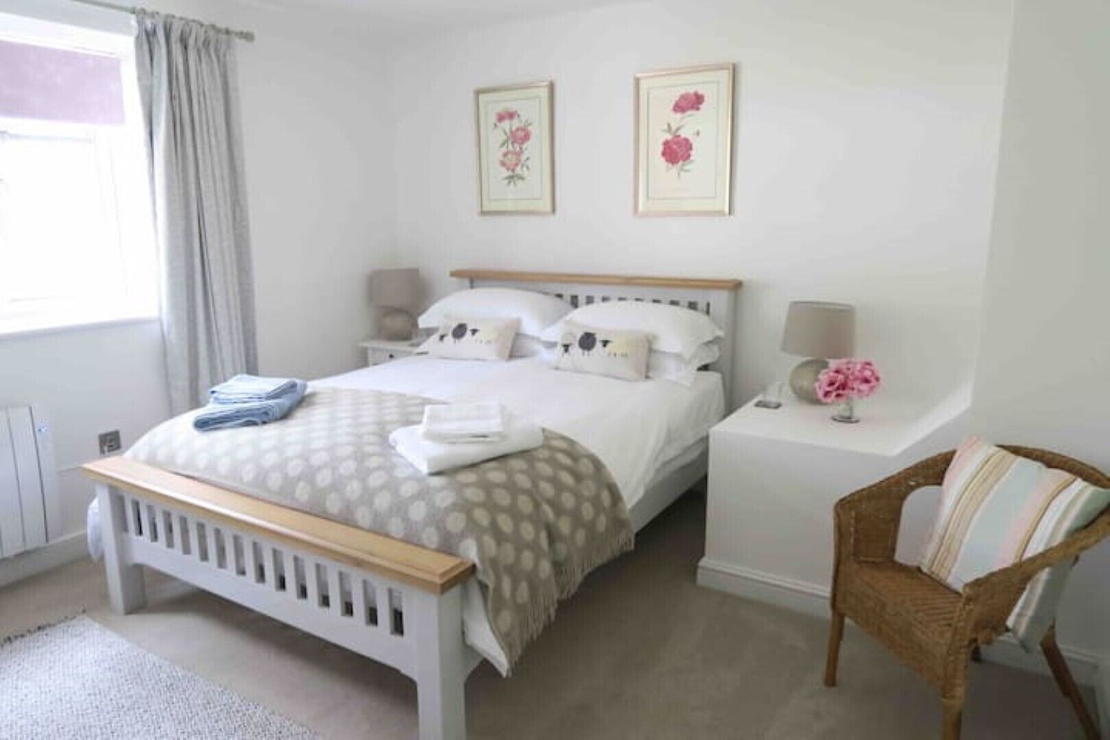 Een keurig opgeruimde slaapkamer met een bed, nachtkastjes, een stoel en bloemrijk kunstwerk in een lijst aan de muur.