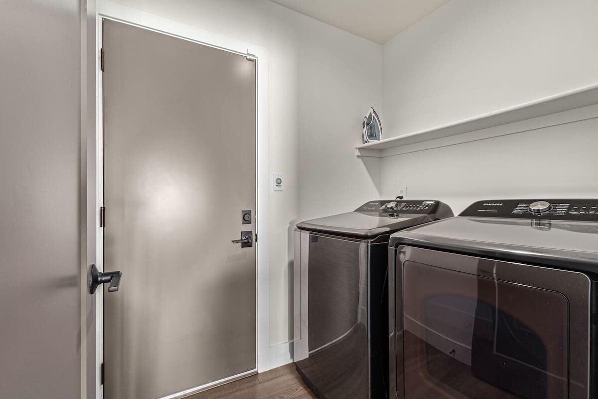 A laundry room with two front-loading washers and dryers, a white door, and a small shelf above the dryers.