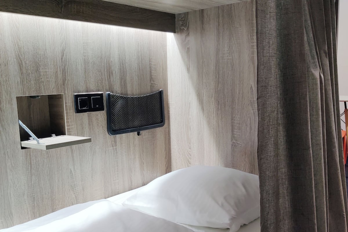 A single bed with white bedding, a small shelf unit, and a wall-mounted air vent.