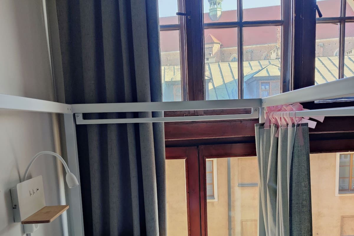 A bunk bed with white bedding, a bedside lamp, and a view of buildings through a window with curtains.