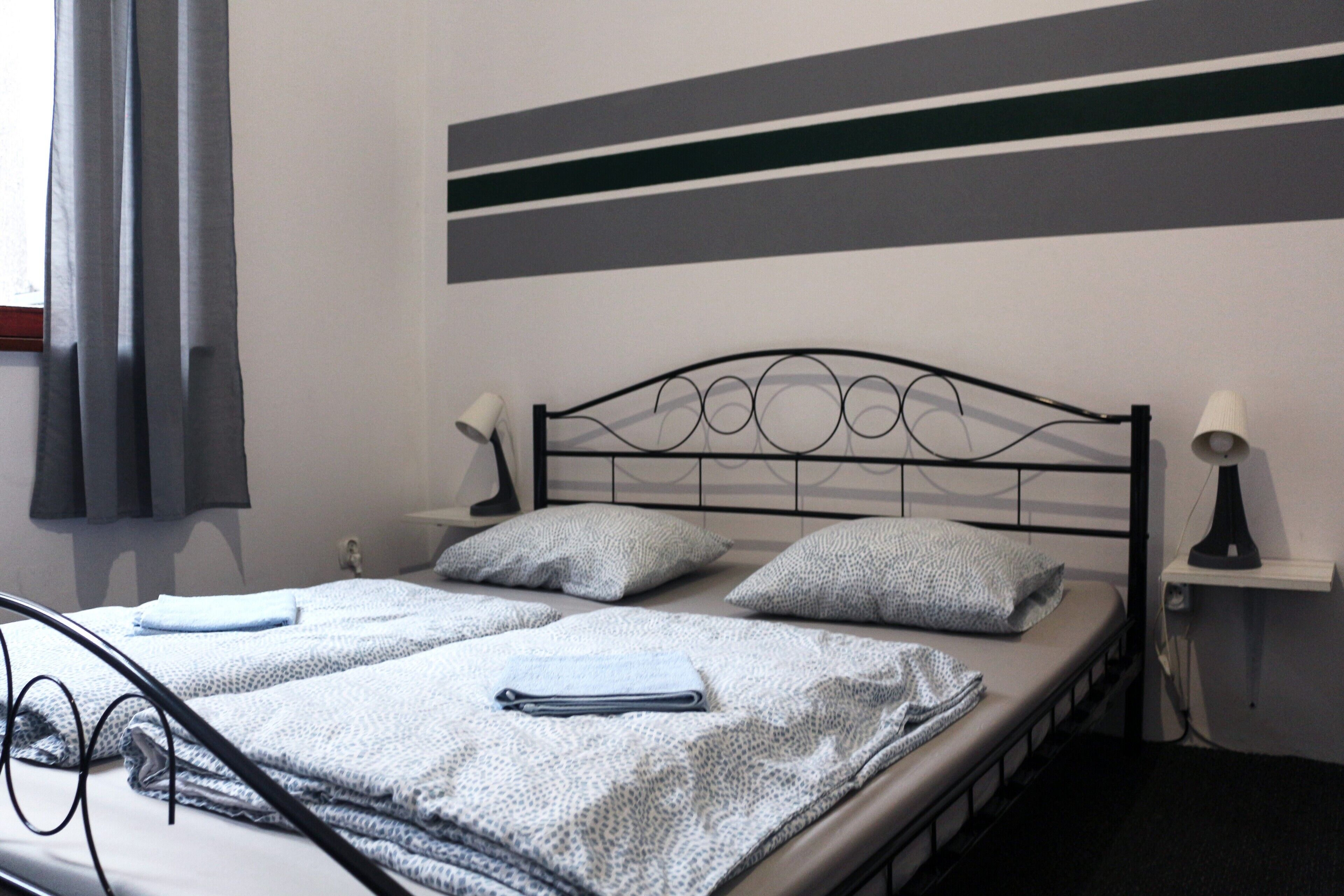 Un lit bien fait, recouvert d’une couvre-lit à motifs, avec deux oreillers et une serviette pliée. Il y a deux lampes de chevet et une petite étagère avec une lampe. La pièce présente un aménagement simple et épuré, avec un mur à rayures au-dessus du lit.