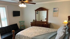 A bedroom with a bed, a dresser, a mirror, a ceiling fan, a TV, and a window with blinds.