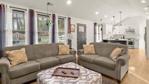 A modern living room with two grey sofas, a patterned ottoman, and a kitchen in the background. - NEW Decatur Bungalow - World Cup Ready Ready (Candler-McAfee)