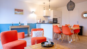 Una sala de estar moderna con mobiliario naranja brillante, una mesa de centro de madera con vasos y un libro, y una zona de cocina al fondo.