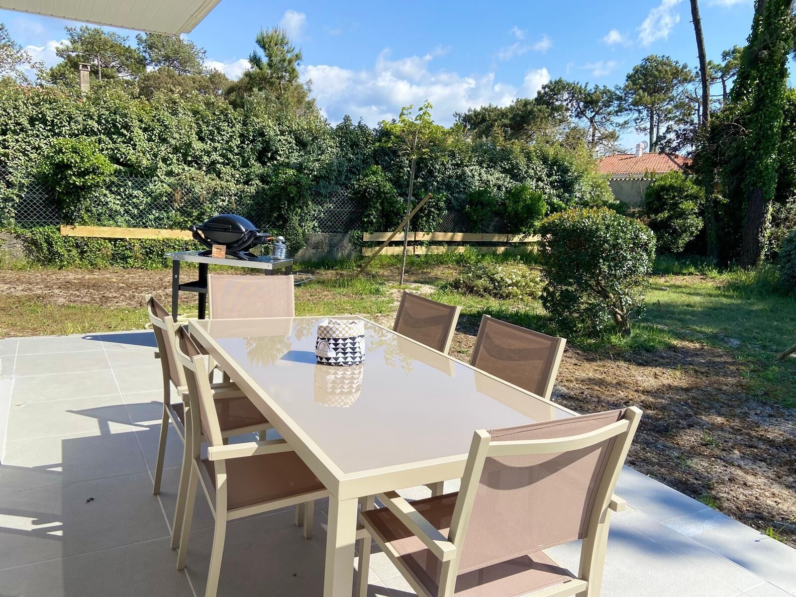 A patio with a table and chairs, surrounded by greenery and a barbecue grill.