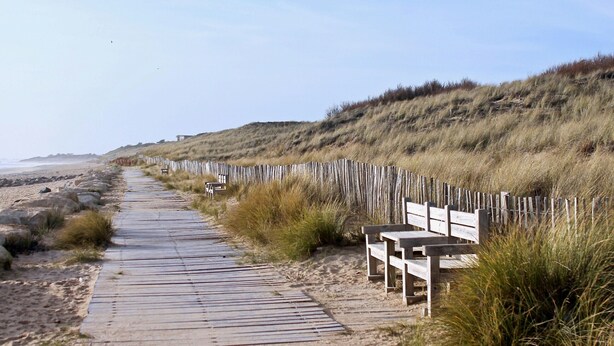 Una passerella in legno che conduce a una spiaggia, con panchine e una recinzione di legno lungo il percorso.