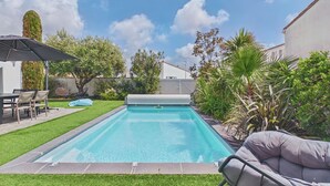 Una piscina rettangolare con una terrazza circostante, un portico coperto con sedie e diverse piante.
