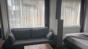 Une pièce avec un canapé gris, une petite table en bois, un lit avec un matelas blanc et une couverture violette, et de grandes fenêtres ornées de rideaux légers.