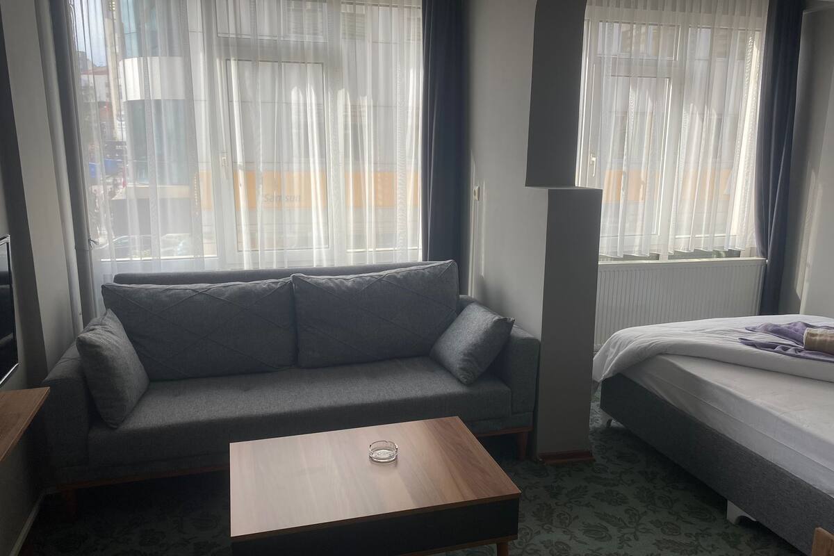 A room with a grey sofa, a small wooden table, a bed with a white mattress and a purple blanket, and large windows with sheer curtains.
