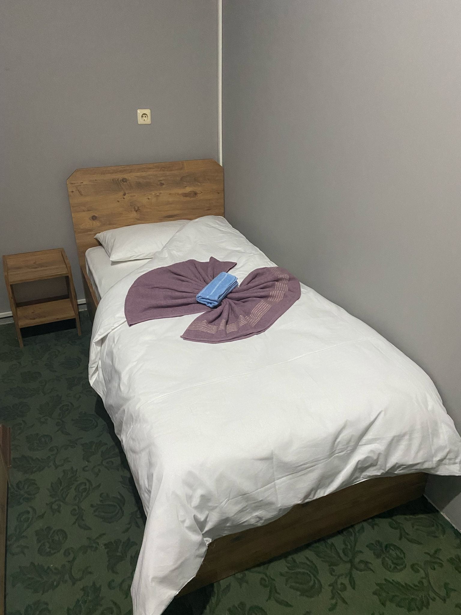Una cama bien hecha con cabecera de madera, una mesita de noche de madera y una alfombra estampada.