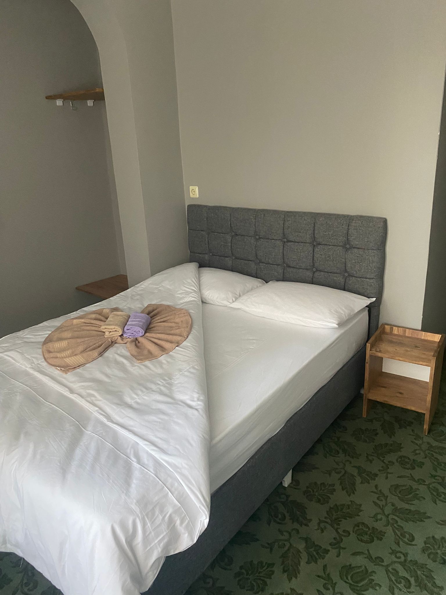 Un lit bien fait, avec une tête de lit grise, une table de chevet en bois et un tapis à motifs verts.