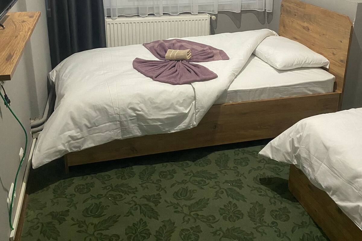 A bedroom with a bed, a radiator, a window with sheer curtains, a wooden headboard, and a green patterned carpet.