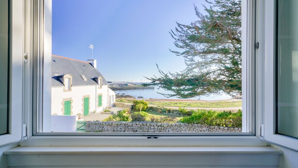 A window with a view of a building with green shutters, a stone wall, and a tree.