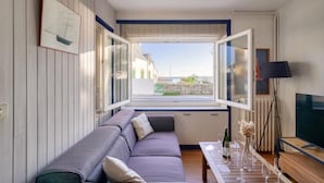 A modern living room with a sofa, a dining table with wine glasses, a TV, and a large window offering a view of the sea.