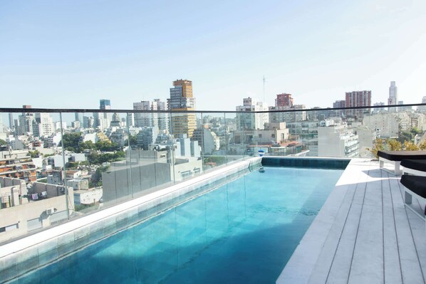 Una piscina en la azotea con vistas a la ciudad, rodeada de barandillas de cristal y suelo de madera.