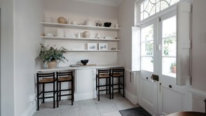 Een keuken met witte kasten, een centraal eiland met stoelen, planken met keukengerei en een raam met uitzicht op groen.