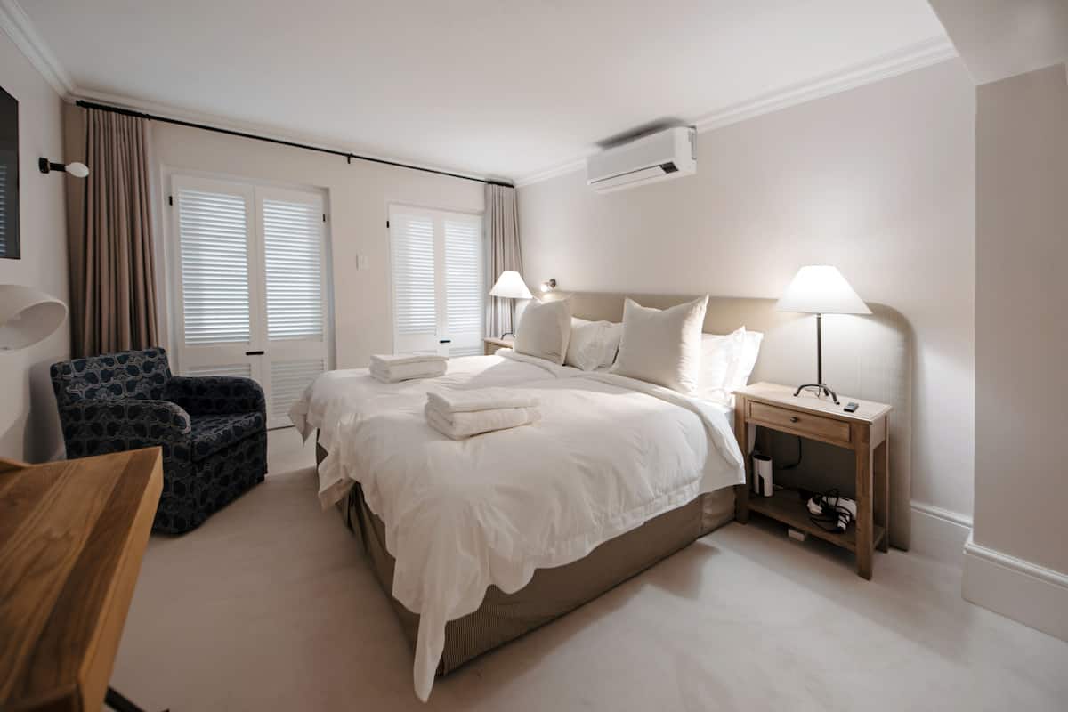 Quarto de hotel com uma cama grande, duas mesas de cabeceira com luminárias, uma cadeira e uma mesa lateral de madeira.