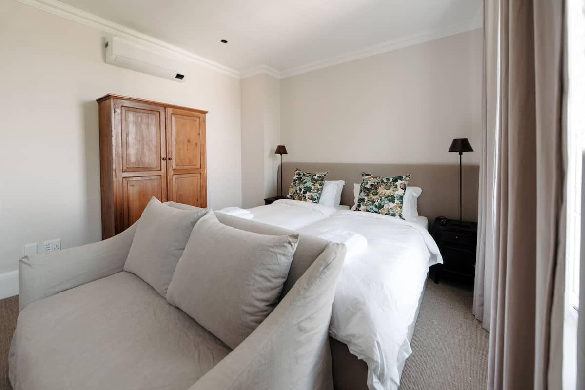 Quarto de hotel com duas camas, um guarda-roupa de madeira, um sofá e uma mesa de cabeceira com abajur.