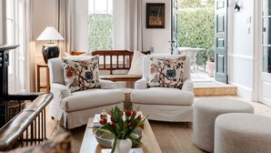 Een ruime woonkamer met een zithoek, een salontafel met bloemen, een lamp en uitzicht op een tuin door grote ramen.