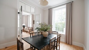 Een eetgedeelte met een donker houten tafel, zes stoelen en een vaas met bloemen. Er is een raam met gordijnen en uitzicht op groen.