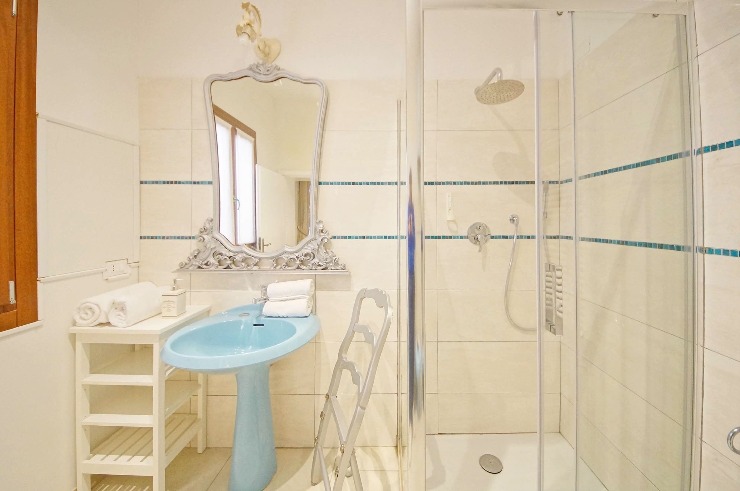 A bathroom with a pedestal sink, a mirror, a shower enclosure, and a white shelf with towels.