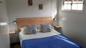 Cama bem arrumada com manta azul e duas almofadas, cabeceira de madeira, mesas de cabeceira com abajures e uma janela com persianas.