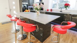 A modern kitchen with a central island, red bar stools, a fruit bowl, and a vase with flowers. - The Cottage at Cromlech Manor Farm (Tyn-y-Gongl)