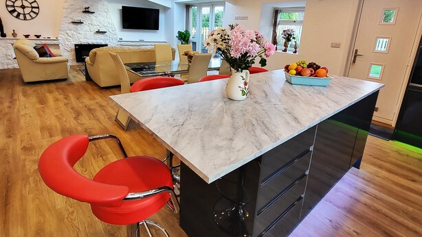 A modern kitchen with a marble countertop, red bar stools, a central island with a vase of flowers, and a dining area with a sofa and chairs. - The Cottage at Cromlech Manor Farm (Tyn-y-Gongl)