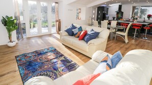 A spacious living room with a large sofa, a coffee table, and a patterned rug. There is a dining area with a kitchen in the background. - The Cottage at Cromlech Manor Farm (Tyn-y-Gongl)