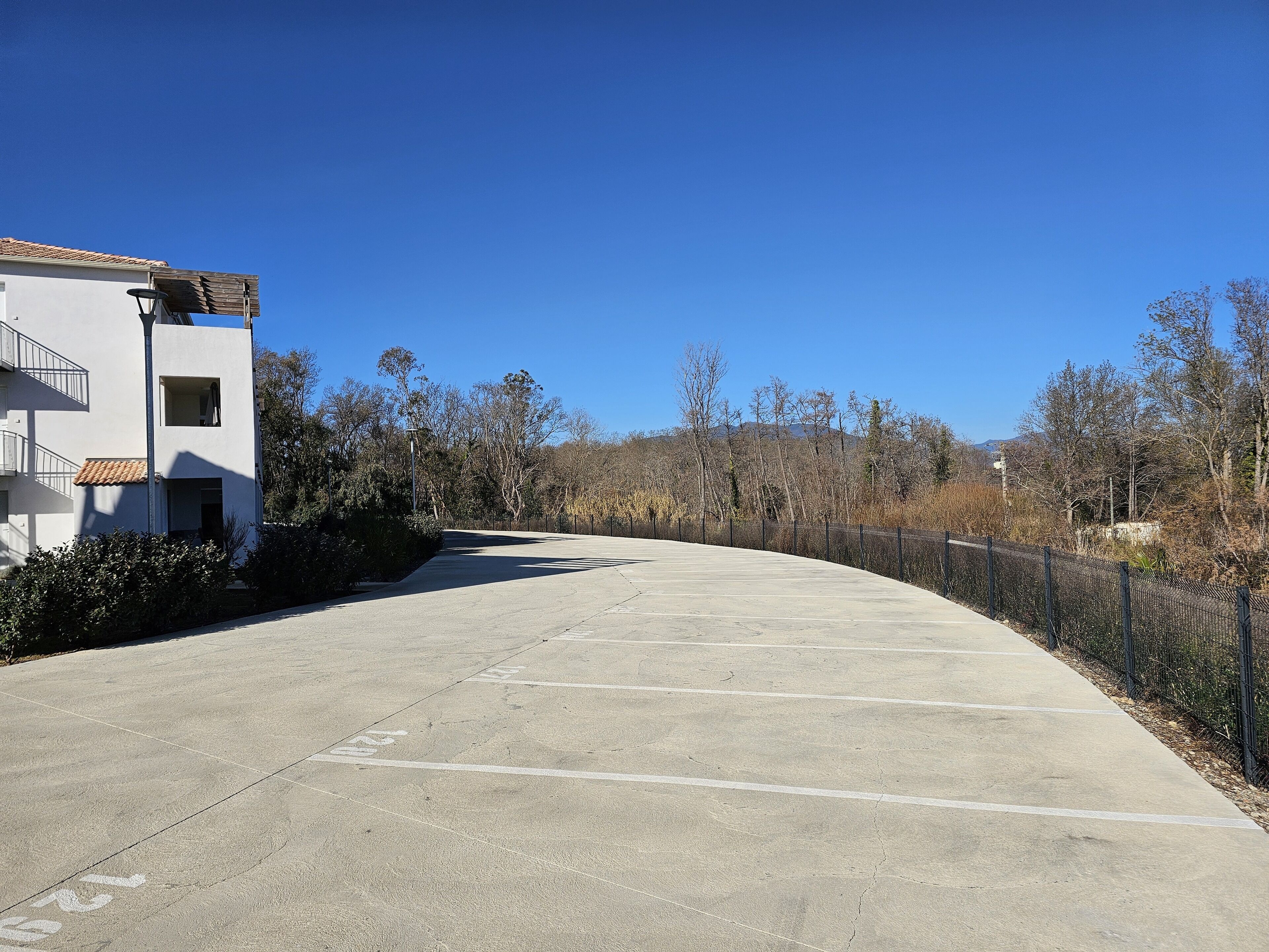 Un parking avec un bâtiment sur la gauche, entouré d’arbres et sous un ciel dégagé.