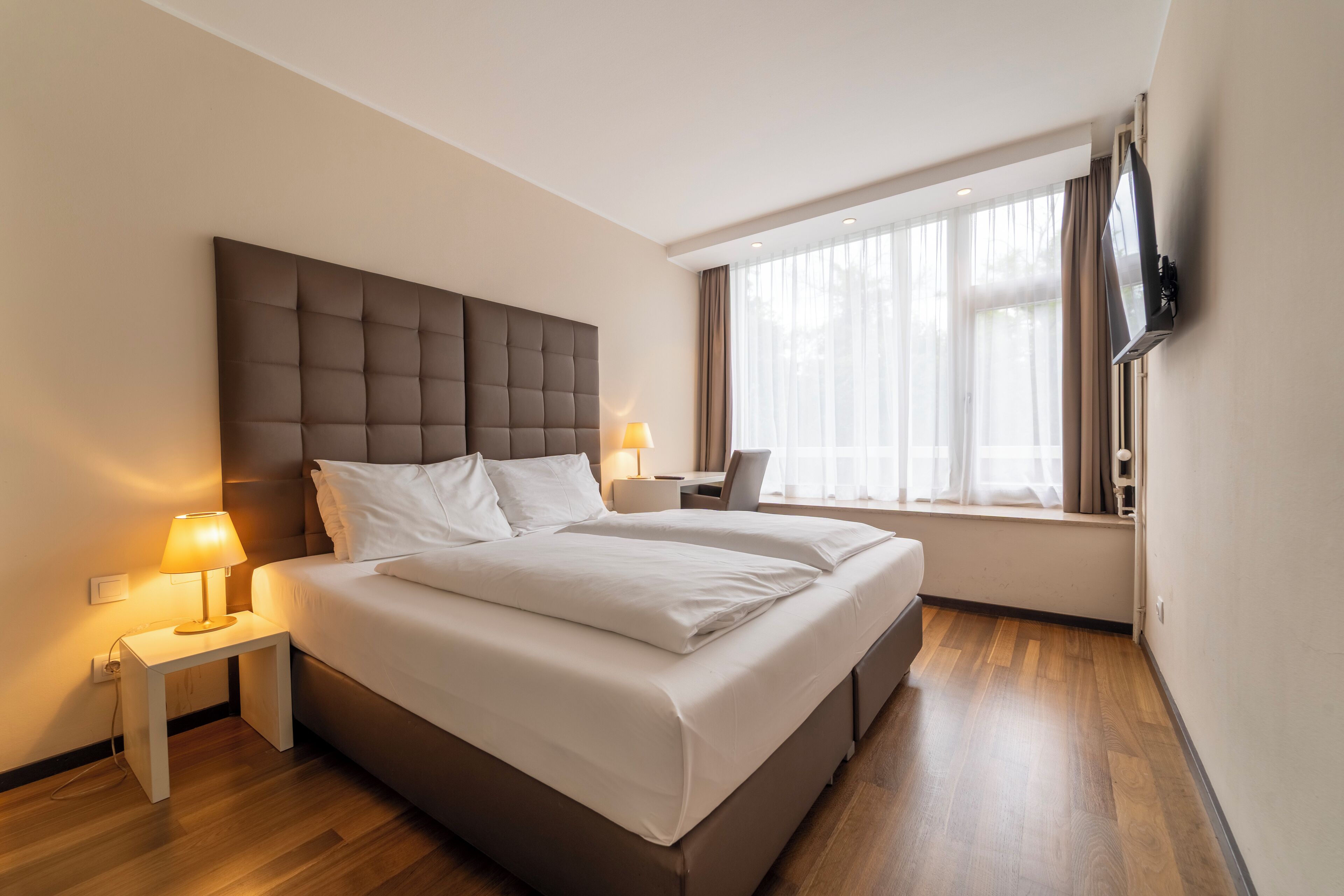Una habitación de hotel moderna con una cama grande, mesitas de noche con lámparas, un televisor de pantalla plana montado en la pared y un ventanal con cortinas.