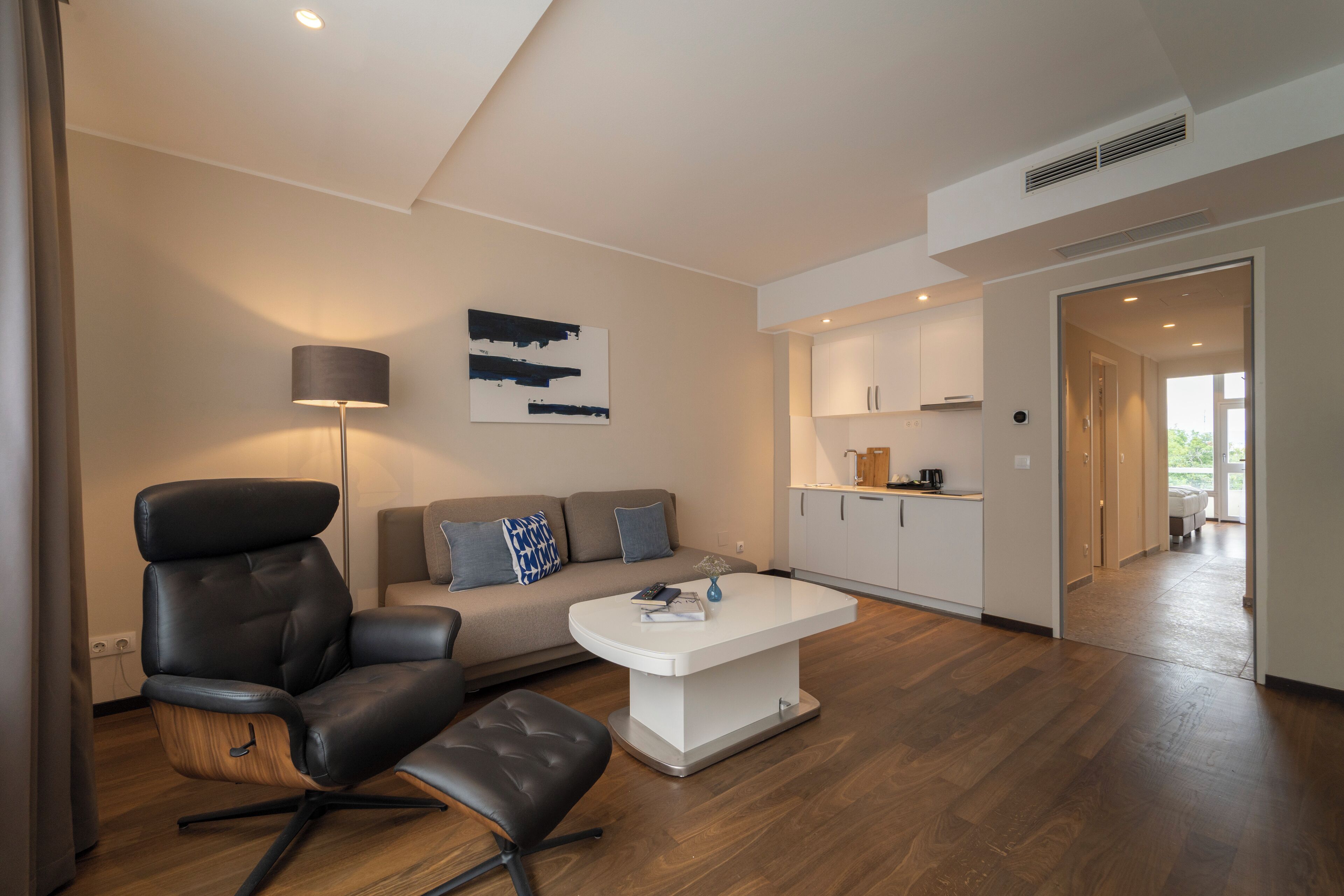 A modern living room with a leather chair, a white coffee table, and a sofa with cushions. There is a painting on the wall and a kitchen area in the background.